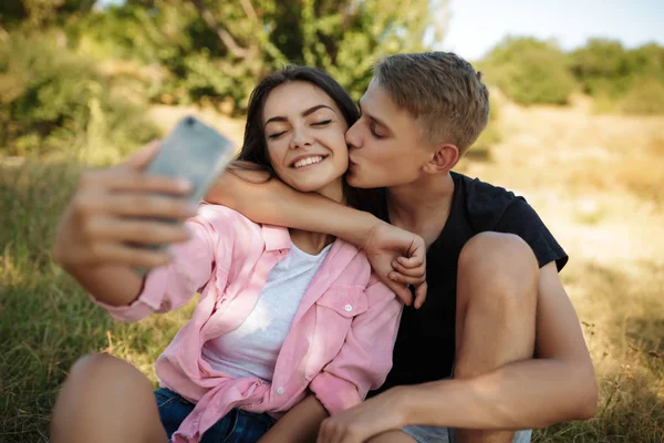 Porträtt av unga leende paret sitter på gräsmattan i parken och att göra selfie. Vackra par att ta foton på frontal mobilkamera — Stockfoto