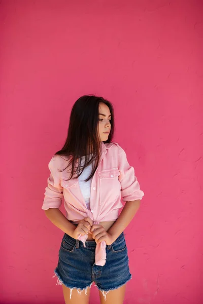 Retrato de menina bonita com cabelos escuros de pé em shorts jeans e camisa enquanto sonhadoramente olhando de lado no fundo rosa isolado — Fotografia de Stock