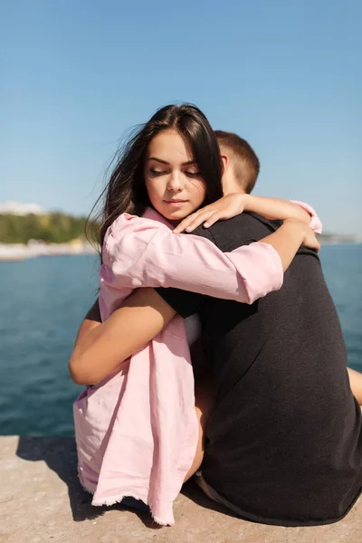 Oturma ve düşünceli bir kenara arka plan üzerinde Deniz Manzaralı bakarken serin çocuk kucaklayan genç güzel kız portresi — Stok fotoğraf