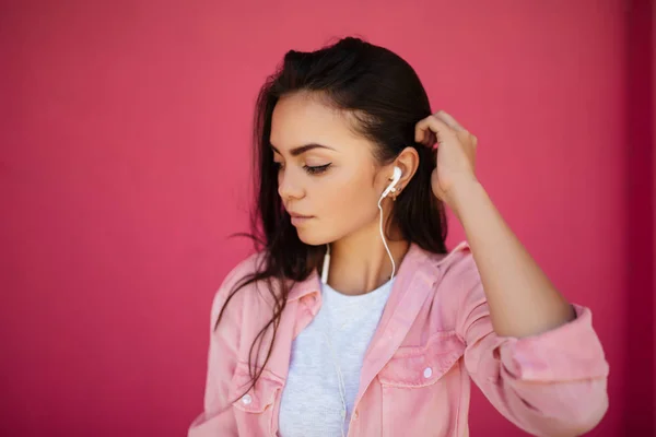 Potret gadis muda cantik dengan rambut hitam berdiri dan mendengarkan musik di earphone sementara memikirkan latar belakang merah muda terisolasi — Stok Foto