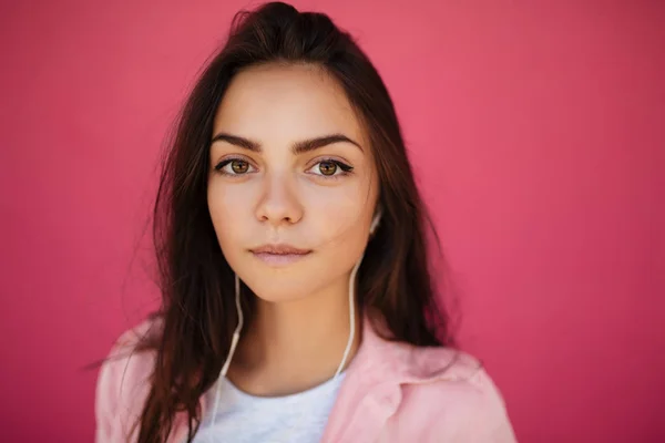 Nahaufnahme Foto von jungen nachdenklichen Mädchen mit dunklen Haaren stehen und Musik hören in Kopfhörer, während verträumt in die Kamera auf rosa Hintergrund isoliert — Stockfoto