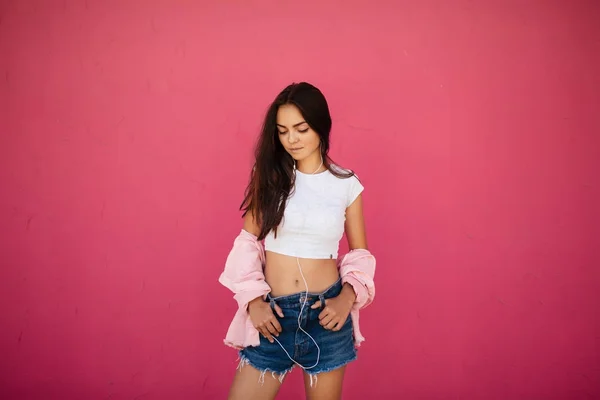 Портрет вдумливої дівчини з темним волоссям, що стоїть в джинсових шортах і білому верхньому куті, слухаючи музику в навушниках на рожевому фоні ізольовано — стокове фото