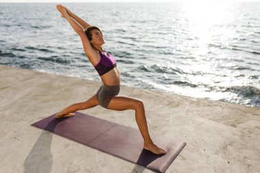 Ayakta ve yoga eğitim genç gülümseyen kadın deniz kenarında teşkil etmektedir. Sportif üst ve yoga uygulamak ve mutlu bir şekilde arka planda muhteşem deniz manzaralı seyir şort güzel Bayan