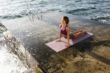 Ellerinin üzerinde yaslanmış ve sersem bir kenara yoga eğitimi sırasında seyir siyah kısa saçlı güzel kadınla deniz kenarında teşkil etmektedir. Sportif üst ve pratik yoga arka plan üzerinde deniz şort gülümseyen kadın