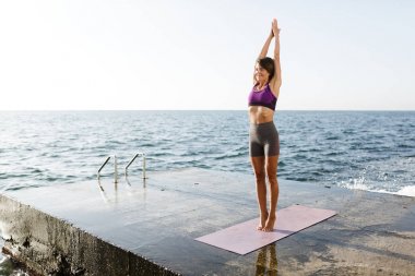 Gülümseyen kadın ayakta ve eğitim yoga pozlar denizin kenarında iken elleri yanaşıyor. Sportif üst ve şort pratik yoga arka plan üzerinde muhteşem deniz manzaralı güzel Bayan