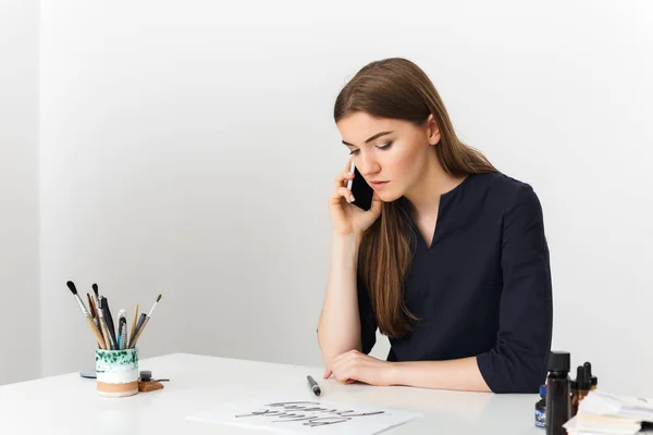 Retrato de jovem senhora agradável sentado na mesa branca e falando em seu celular enquanto sonhadamente olhando para papel isolado — Fotografia de Stock