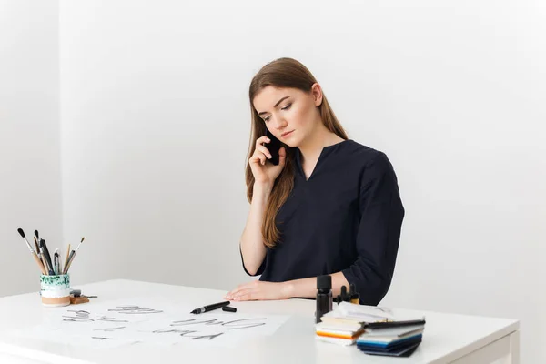 Porträt einer jungen hübschen Dame, die am weißen Schreibtisch sitzt und mit ihrem Handy spricht, während sie nachdenklich zur Seite schaut — Stockfoto