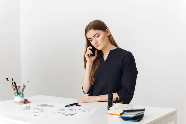 Retrato de jovem bonita senhora sentada na mesa branca e falando em seu celular, enquanto pensativamente olhando para o lado isolado — Fotografia de Stock