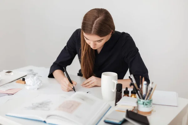 Portré, fiatal, gyönyörű hölgy ül a fehér íróasztal és írás megjegyzi a papír elszigetelt nyitott könyv — Stock Fotó