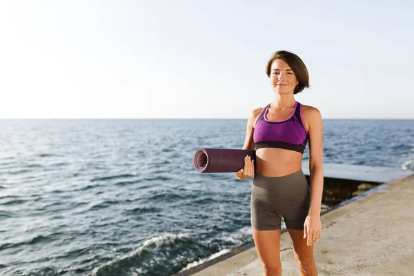 Retrato de una joven sonriente con el pelo corto oscuro sosteniendo la esterilla de yoga en la mano mientras camina junto al mar. Niza dama en top deportivo y pantalones cortos de pie y mirando felizmente en la cámara con el mar en el fondo —  Fotos de Stock