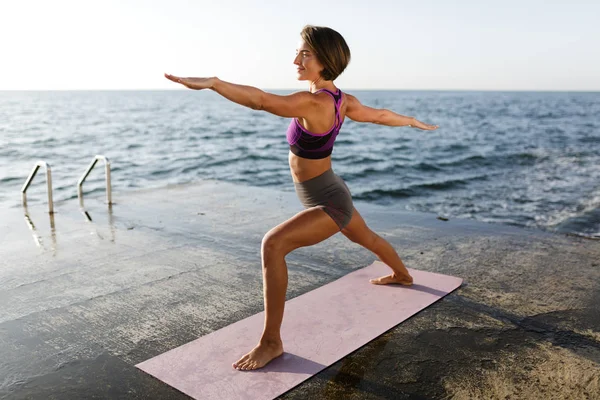 Портрет женщины с темными короткими волосами, стоящей и оттягивающей руки в сторону во время тренировки йоги позы у моря. Девушка в спортивном топе и шорты практикующие йогу с морем на заднем плане — стоковое фото
