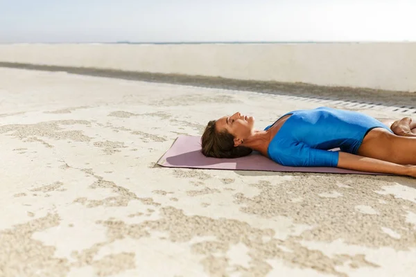 Young woman with dark sort hair lying on yoga mat and training yoga poses while dreamily closing her eyes. Portrait of beautiful lady in blue swimsuit meditating and practicing yoga isolated