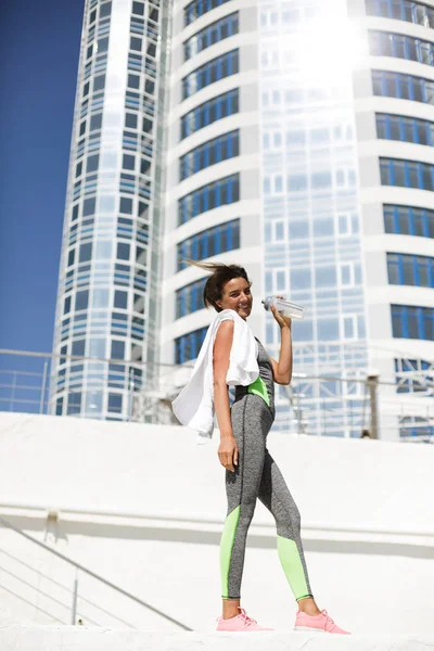 Portrét radostné ženy v moderní šedé sportovní oblek a růžové tenisky stojící s bílým ručníkem na rameni a šťastně pohledu kamery s vysokou budovou na pozadí — Stock fotografie