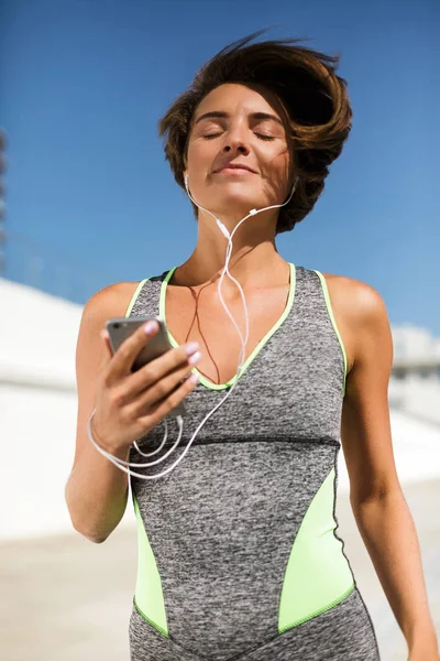 Portrait of beautiful smiling woman with brown short hair in modern gray sport suit holding cellphone with earphones and dreamily closing her eyes while jogging isolated