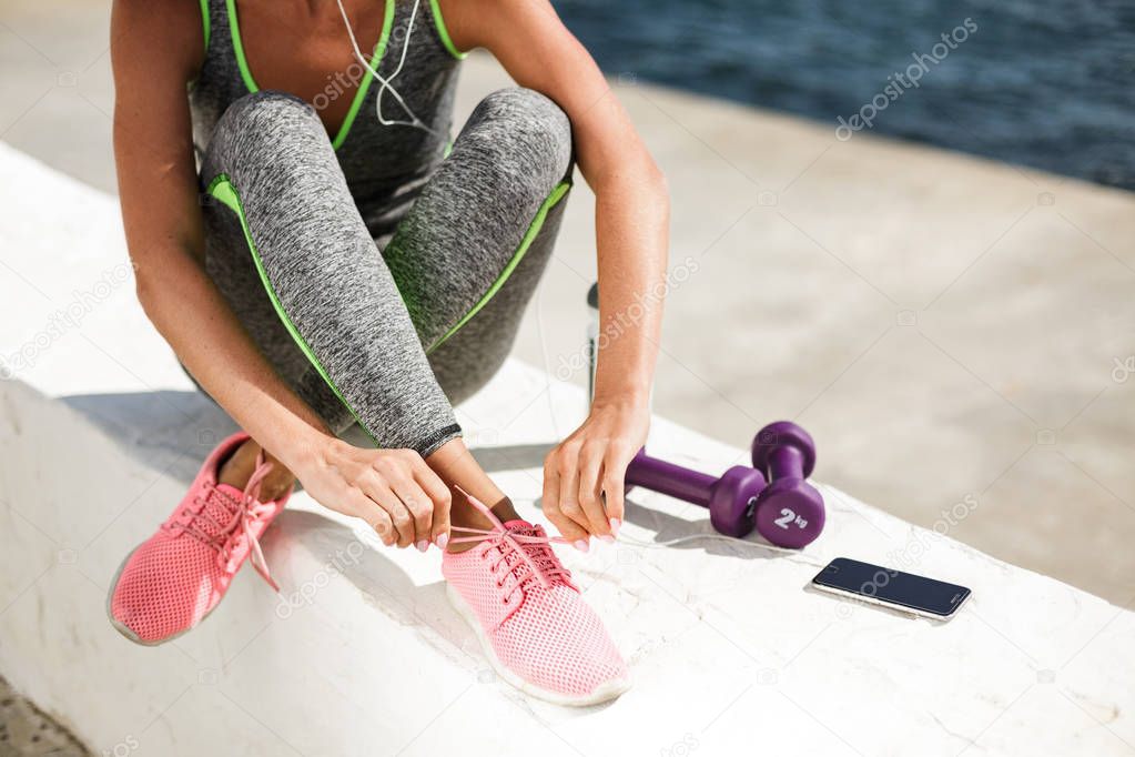 Portrait of woman hands and legs in pink sneakers sitting and tie shoelaces with purple dumbbells,cellphone and bottle of water nearby