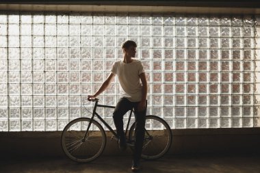 Klasik bisiklet ile ayakta ve sersem bir kenara kameraya poz süre seyir serin çocuk. Beyaz t-shirt bisiklet ile arka plan cam duvara yaslanmış genç düşünceli adam