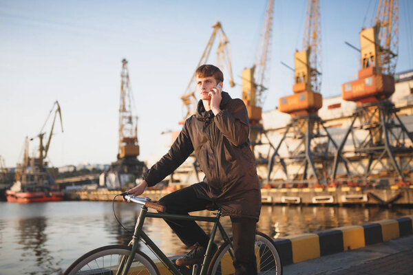 Portrait of young man with brown hair standing with bicycle and thoughtfully looking aside while talking on his mobile phone. Cool boy in down jacket on bicycle standing with sea port on background