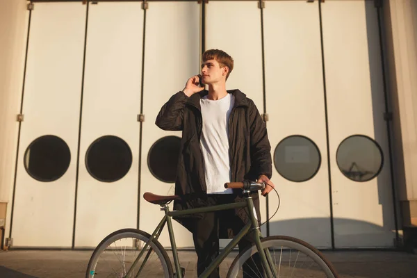 Young man with brown hair standing with classic bicycle and dreamily looking aside while talking on his cellphone. Portrait of boy in white t-shirt and down jacket standing with bicycle on street