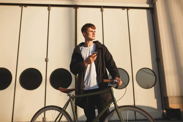 Portrait of young man with brown hair standing with classic bicycle and dreamily looking aside with cellphone in his hand. Smiling boy in down jacket walking with bicycle along city streets — Stock Photo, Image