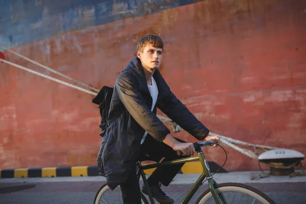 Retrato de un hombre joven con cabello castaño montando bicicleta clásica mientras mira a un lado de ensueño. Niño reflexivo en chaqueta y mochila negra montar en bicicleta — Foto de Stock