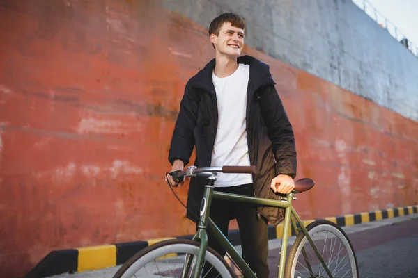 Retrato de un joven de cabello castaño caminando con una bicicleta clásica mientras mira alegremente a un lado. Chico sonriente en chaqueta y mochila negra de pie con bicicleta en la calle — Foto de Stock