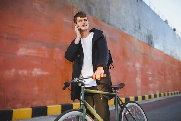 Retrato de un joven de cabello castaño caminando con una bicicleta clásica y mirando felizmente a un lado mientras habla por su teléfono celular. Chico sonriente en chaqueta y mochila negra de pie con bicicleta — Foto de Stock