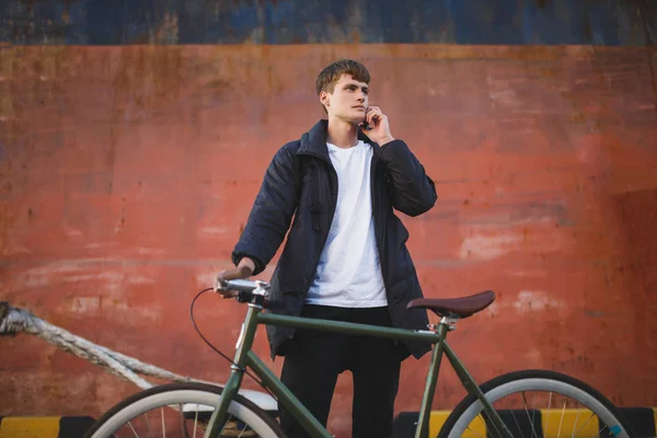 Thoughtful boy with brown hair standing with bicycle and dreamily looking aside while talking on his cellphone. Young man in down jacket standing with bicycle and mobile phone in hand — Stock Photo, Image