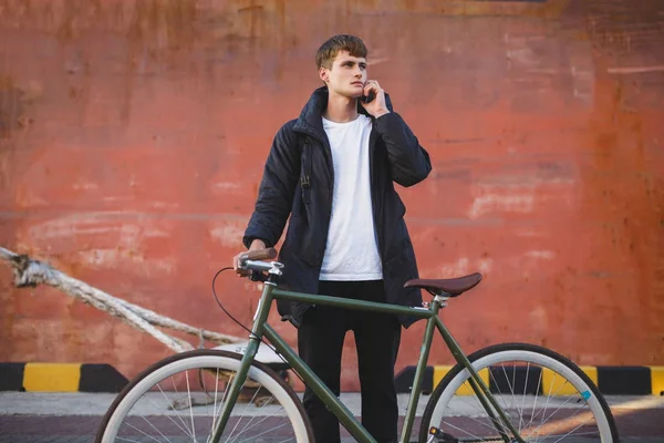 Portrait of boy with brown hair standing with classic bicycle and thoughtfully looking aside while talking on his cellphone. Young man in down jacket standing with bicycle — Stock Photo, Image