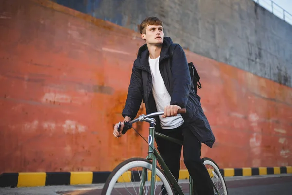 Thoughtful boy with brown hair riding bicycle while dreamily looking aside. Portrait of young man in down jacket and black backpack riding bicycle on street — Stock Photo, Image