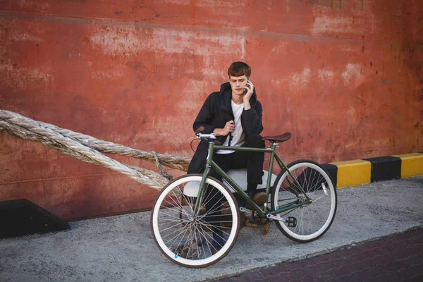 Cool boy with brown hair sitting and dreamily looking aside while talking on his cellphone. Young thoughtful man in down jacket and white t-shirt sitting with mobile phone in hand and bicycle nearby — Stock Photo, Image