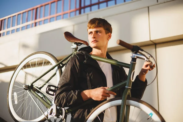 Portrait of thoughtful boy with brown hair standing and holding bicycle while dreamily looking aside. Young man in down jacket standing with classic beautiful bicycle on street — Stock Photo, Image