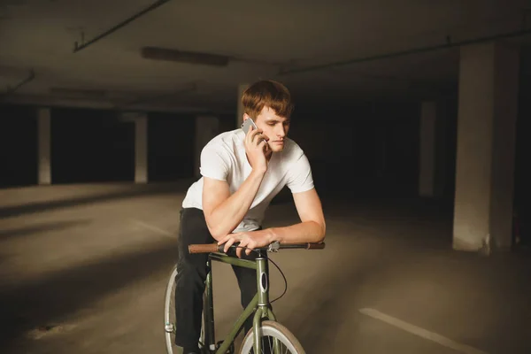 Portrét mladíka zamyšleně sedí na kole a mluví na jeho mobilní telefon. Cool chlapce s hnědými vlasy v bílé tričko, opřel se o kolo při používání mobilního telefonu — Stock fotografie