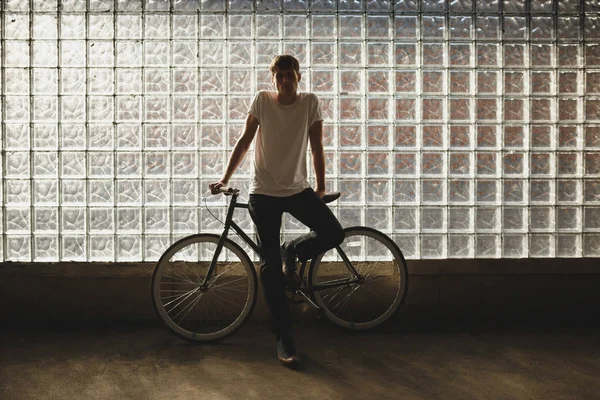 Băiat cool în picioare cu bicicleta clasică și atent în cameră. Fotografie a unui tânăr în tricou alb sprijinit pe bicicletă cu perete de sticlă pe fundal — Fotografie, imagine de stoc