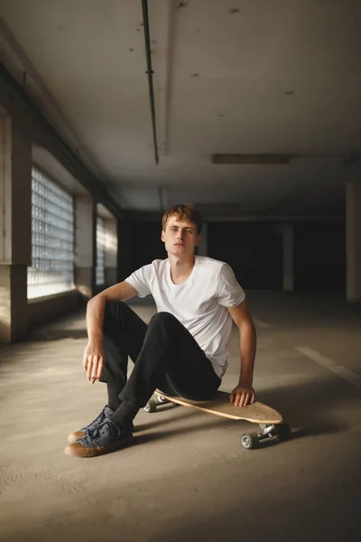 Portrait of cool boy with brown hair sitting on skateboard and dreamily looking in camera. Young thoughtful man in white t-shirt sitting and holding cigarette in hand while posing on camera — Stock Photo, Image