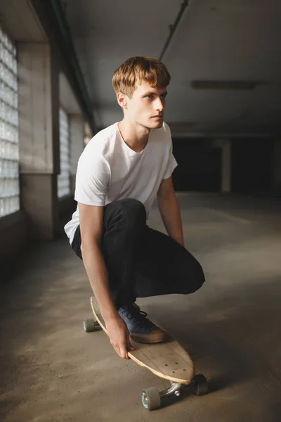 Portrait of young man with brown hair standing on skateboard and dreamily looking aside while posing on camera. Young thoughtful man in white t-shirt riding on skateboard — Stock Photo, Image