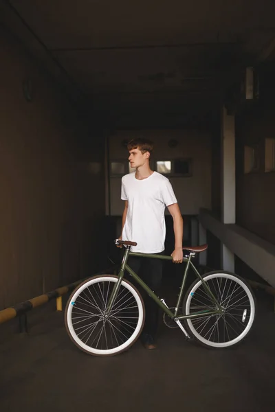 Portret de băiat cu păr castaniu în picioare cu bicicleta și se uită visător deoparte. Fotografie a tânărului om grijuliu în tricou alb în picioare cu bicicleta clasică în parcare — Fotografie, imagine de stoc
