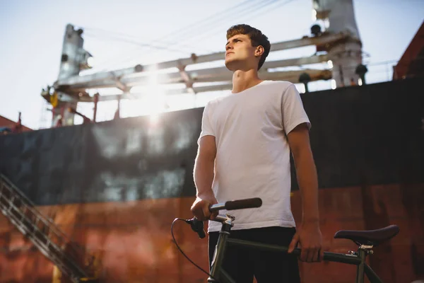 Portrait of serious boy with brown hair standing with bicycle and dreamily looking aside. Young man in white t-shirt standing and posing on camera with big ship on background — Stock Photo, Image