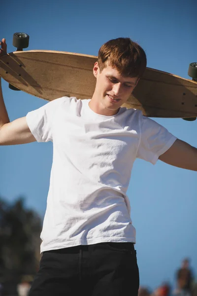 Portrait of young man with brown hair standing with skateboard on his shoulder and dreamily looking down. Young smiling man in white t-shirt standing with skateboard in hand — Stock Photo, Image