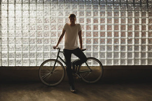 Cool boy standing with classic bicycle and thoughtfully looking in camera. Photo of young man in white t-shirt leaning on bicycle with glass wall on background — Stock Photo, Image