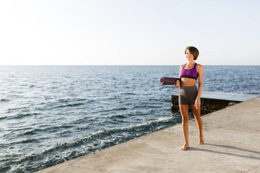 Yoga mat deniz kenarında yürürken elinde tutan siyah kısa saçlı genç gülümseyen kadın. Sportif üst ve ayakta ve sersem bir kenara arka plan üzerinde deniz manzaralı seyir şort güzel Bayan