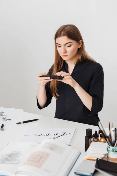 Retrato de jovem bela senhora sentada na mesa branca e tirando fotos de notas legais em seu celular isolado — Fotografia de Stock