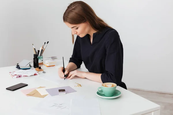 Portré, fiatal, gyönyörű hölgy ül a fehér íróasztal csésze kávé és elszigetelt papír írás — Stock Fotó