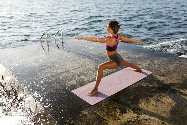 Gülümseyen kadın siyah kısa saçlı ayakta ve eller eğitim yoga pozlar denizin kenarında ise bir kenara çekerek. Sportif üst ve pratik yoga arka plan üzerinde muhteşem deniz manzaralı şort genç bayan
