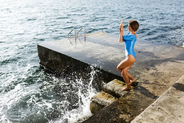 Lächelnde Frau, die am Meer steht und verschiedene Yoga-Posen trainiert. schöne Dame im Badeanzug praktiziert Yoga mit herrlichem Meerblick auf dem Hintergrund — Stockfoto