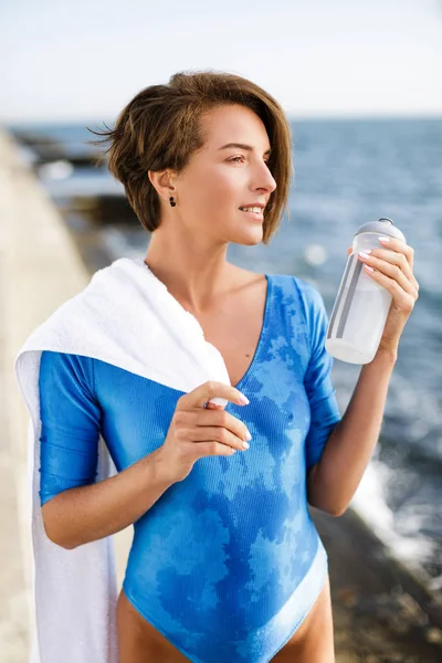 Portret pięknej kobiety w stojący strój kąpielowy niebieski biały ręcznik na ramiona i butelką w ręku sennie patrząc na bok. Miła Pani z stojący ciemne, krótkie włosy, z widokiem na morze na tle — Zdjęcie stockowe