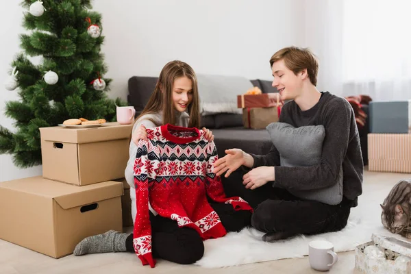 Schönes Mädchen mit neuem Pullover, während es mit dem Jungen auf dem Boden zu Hause sitzt, mit Weihnachtsbaum im Hintergrund — Stockfoto