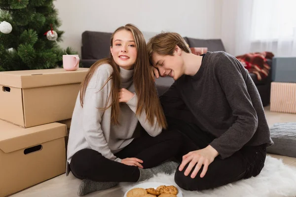 Retrato de una hermosa chica sentada en el suelo con un chico que apoyaba la cabeza sobre su hombro en casa y miraba felizmente en la cámara — Foto de Stock