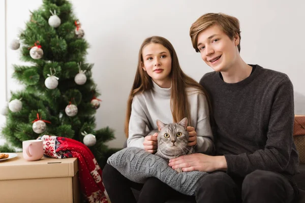 Porträt eines hübschen jungen Paares auf Sofa sitzend mit schöner Katze auf Kissen zu Hause und Weihnachtsbaum in der Nähe — Stockfoto