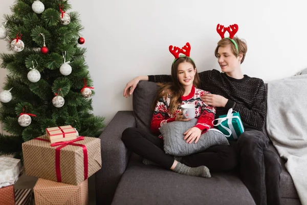Evde kanepede oturan ve geyik boynuzları ile birlikte Noel ağacının yanında vakit iken giyen genç güzel Çift — Stok fotoğraf
