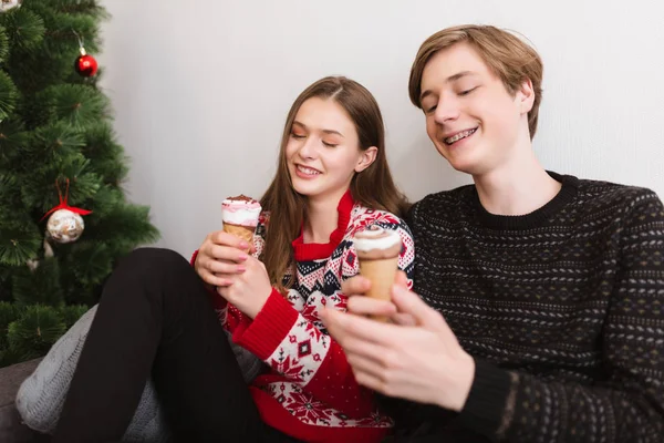 Retrato de una hermosa pareja joven sentada en el sofá en casa y felizmente pasando tiempo juntos mientras comen helado con árbol de Navidad cerca — Foto de Stock